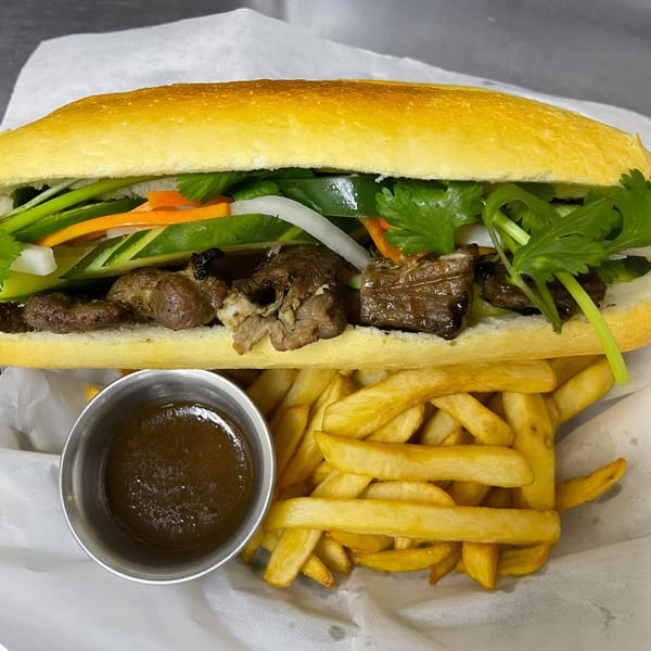 A Vietnamese banh mi sandwich with grilled meat and vegetables, served with fries and dipping sauce.